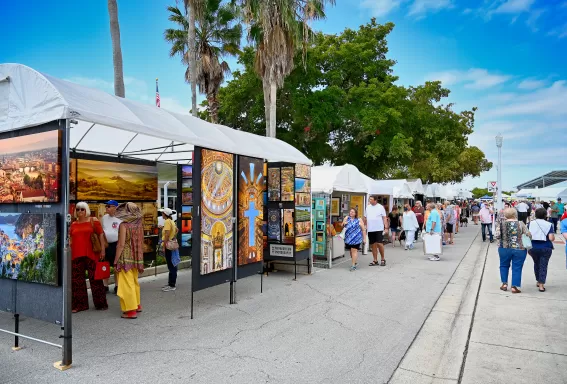 Visitors stand at an outdoor art fair booth examining framed watercolor paintings of birds and coastal wildlife displayed on a black gallery wall, with tents and other attendees visible in the background.